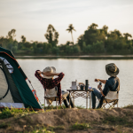 Rear view of young backpacker couple sitting to relax at front of the tent near the lake with coffee set and making fresh coffee grinder while camping trip on summer vacation