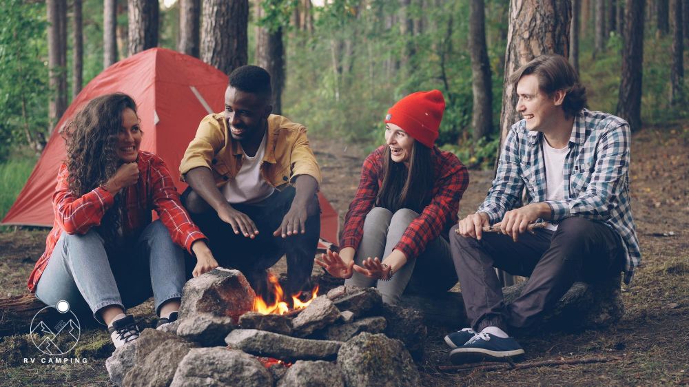 Young men and women travelers are sitting around fire telling camping jokes and  stories. laughing handsome guy is throwing firewood in flame tent and backpacks are visible
